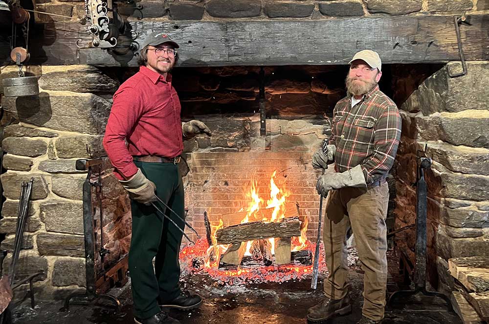 Two Salem Cross Innkeepers smiling in front of the Roasting Jack as they prep the fire.